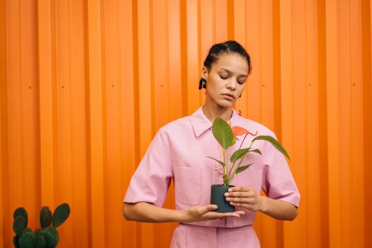 Stylish woman in pink dress holding a potted plant against a vibrant orange background.
