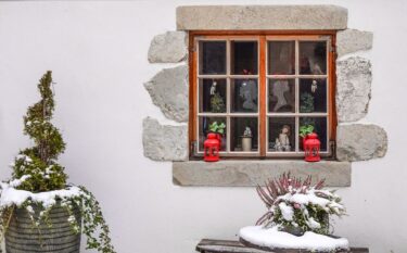 A cozy winter scene featuring a rustic window with red lanterns, ornaments, and snow-covered plants.
