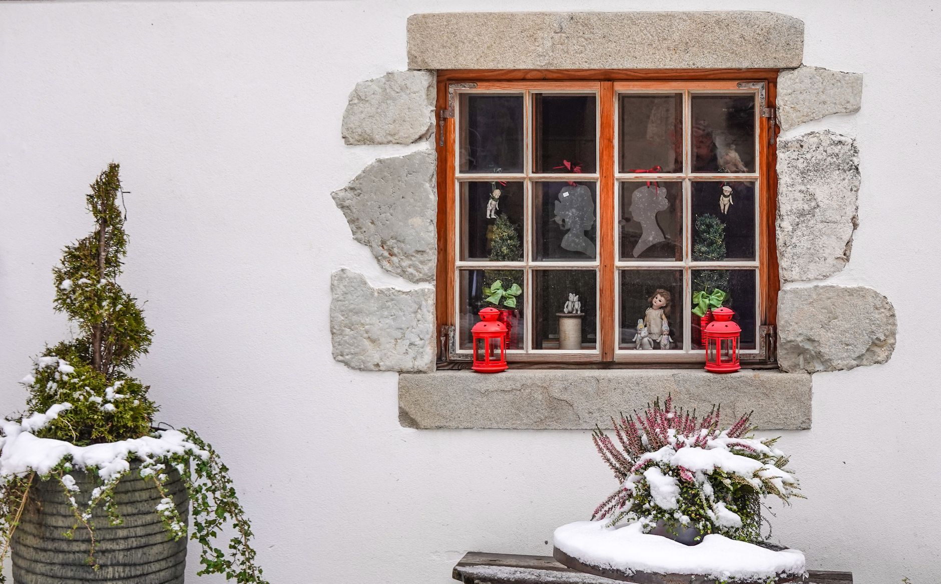 A cozy winter scene featuring a rustic window with red lanterns, ornaments, and snow-covered plants.